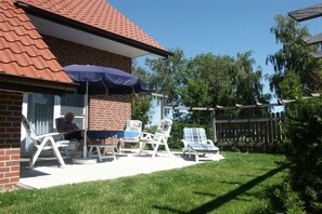 A brick house with a red roof, a covered patio with white lounge chairs, and a wooden fence. - Holiday home with 2 bedrooms (Kaltenhof)