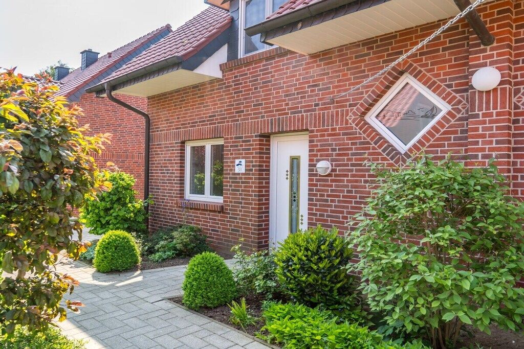 Een bakstenen huis met een witte deur, een raam en een kleine tuin met struiken.