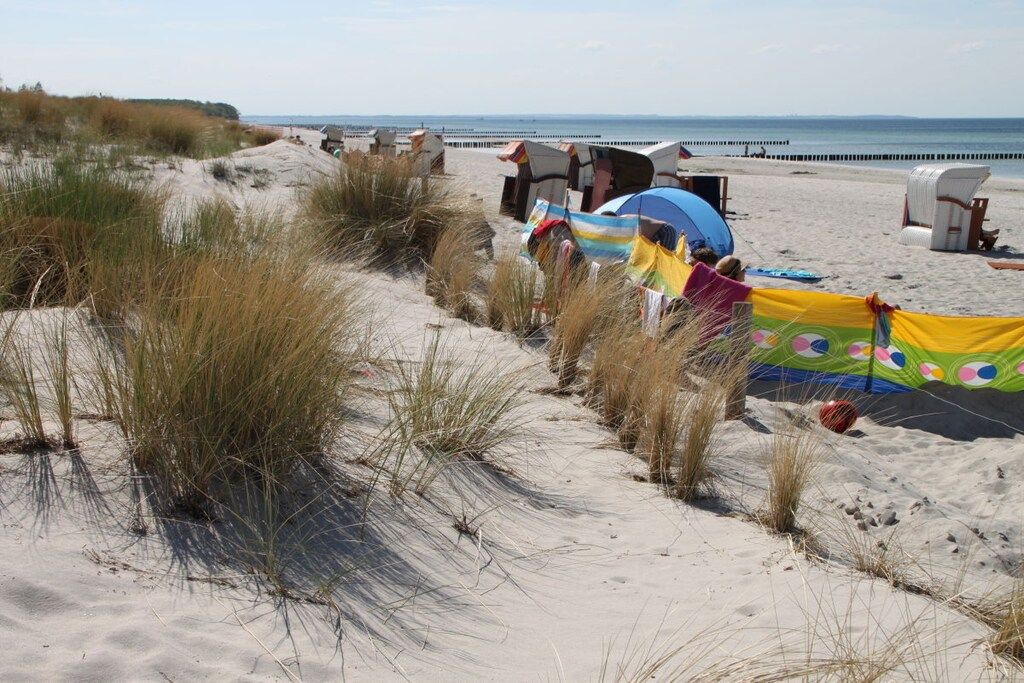 Une plage de sable avec des tentes et des chaises de plage, une jetée en arrière-plan et des dunes couvertes d’herbe.