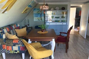 Ein Essbereich mit einem Holztisch, zwei gelben Stühlen, einer farbenfrohen Couch und einer Küche im Hintergrund.