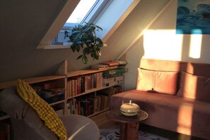 A cozy room with a sloped ceiling, a bookshelf, a cushioned chair, a small round table, and a potted plant. - Holiday apartment with 1 bedroom (Fehmarn)