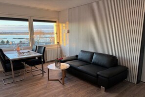 Ein modernes Wohnzimmer mit einem schwarzen Ledersofa, einem Esstisch mit Stühlen, einem runden Couchtisch und Blick auf ein Gewässer durch große Fenster.