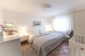 A hotel room with a bed, bedside tables, a lamp, a crib, and a window with curtains. - Holiday apartment with 1 bedroom (Zingst)