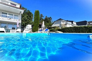 A swimming pool with clear blue water, surrounded by lounge chairs and umbrellas, with residential buildings in the background. - Holiday apartment with 3 bedrooms (Sierksdorf)
