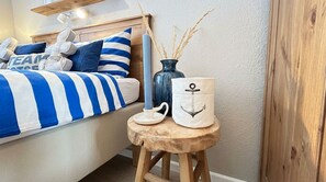 Un dormitorio con una cama con ropa de cama azul y blanca a rayas, un cabecero de madera y un taburete de madera pequeño con objetos decorativos.