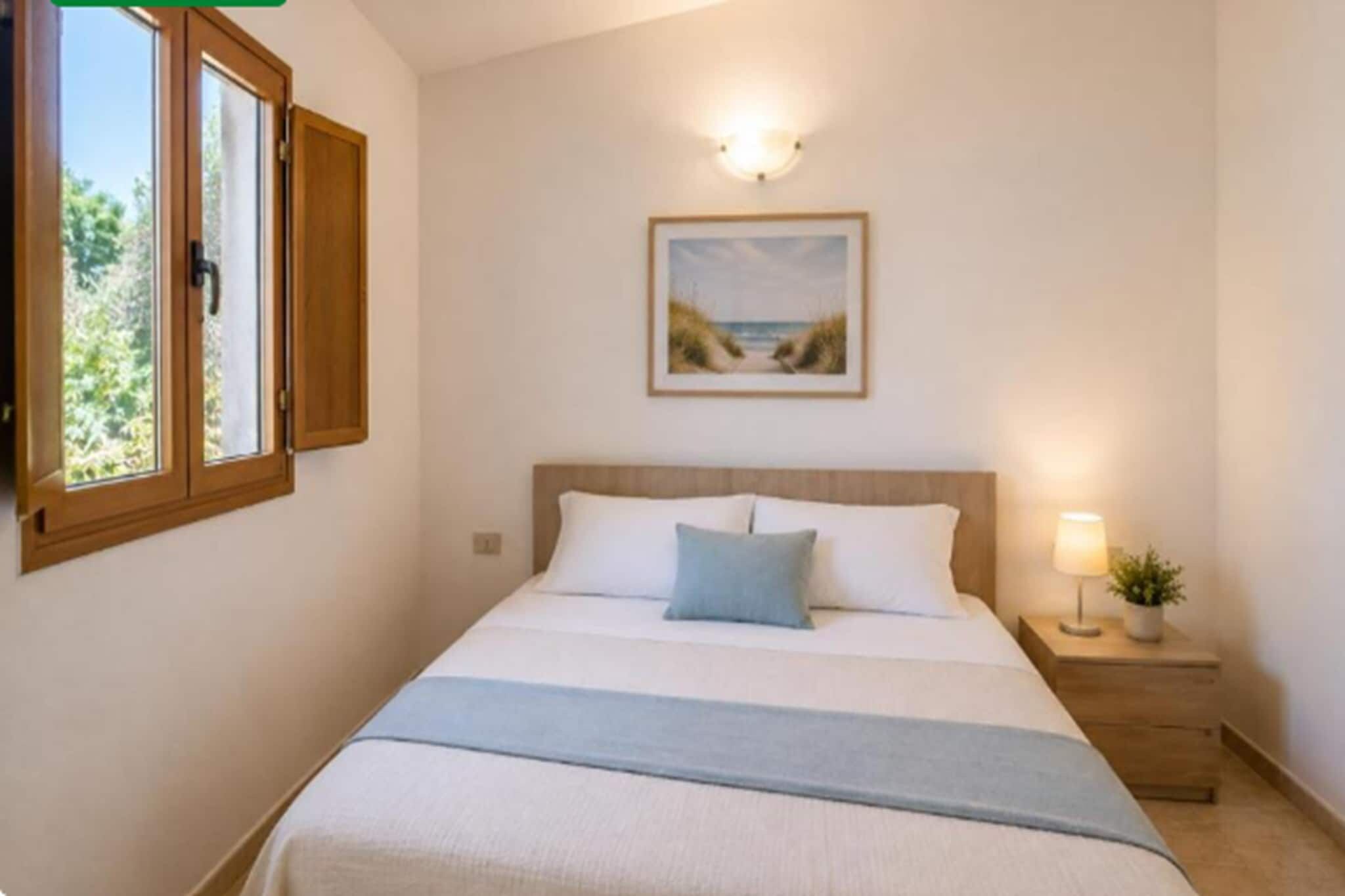 Un dormitorio con cama, mesita de noche, lámpara, ventana con cortinas y un cuadro en la pared.