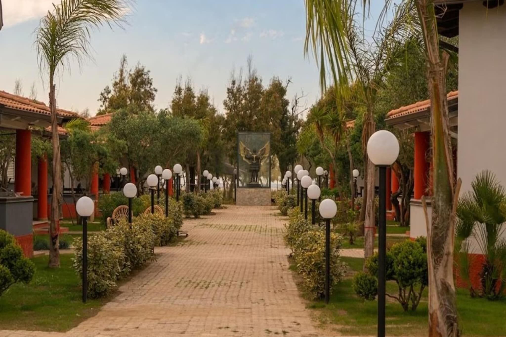 Un vialetto lastricato conduce a un edificio con una statua centrale, circondato da vegetazione e palme.