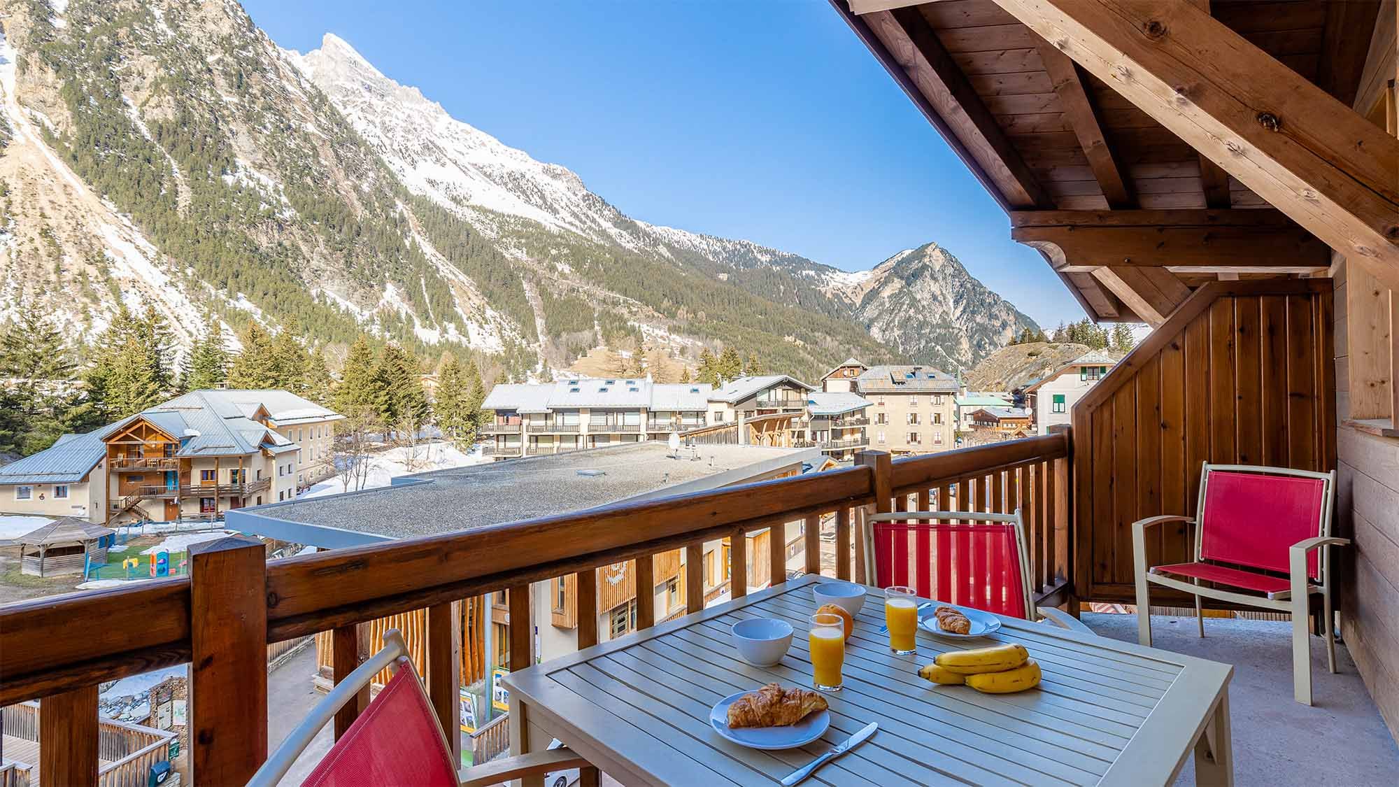 Ein Balkon mit Frühstückstisch, mit Blick auf ein dörfliches Bergdorf mit Gebäuden und schneebedeckten Gipfeln.