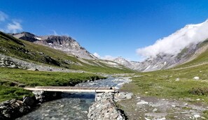 Eine Berglandschaft mit einer Holzbrücke über einem Bach, felsigem Gelände und schneebedeckten Gipfeln.