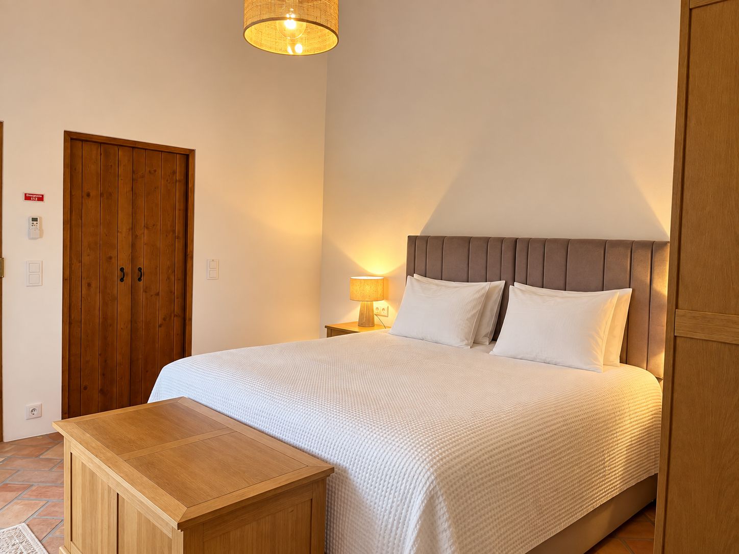 A hotel room with a bed, bedside lamp, wooden wardrobe, and a small wooden table.