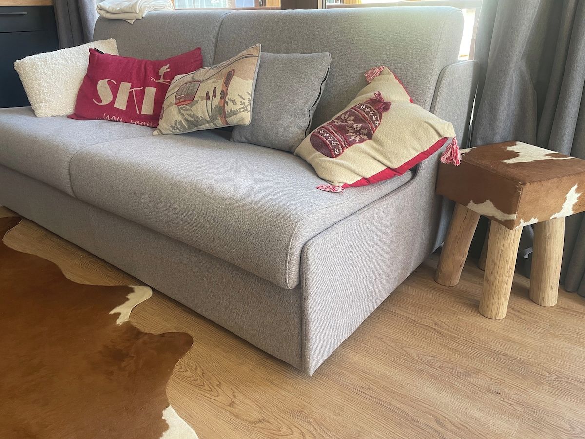 Ein graues Sofa mit gemusterten Kissen, ein Holzschemel und ein Kuhfellteppich in einem Raum mit Holzboden.