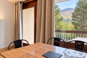Ein Balkon mit Holztisch und -stühlen, Blick auf eine Berglandschaft und eine Tür mit Vorhängen.