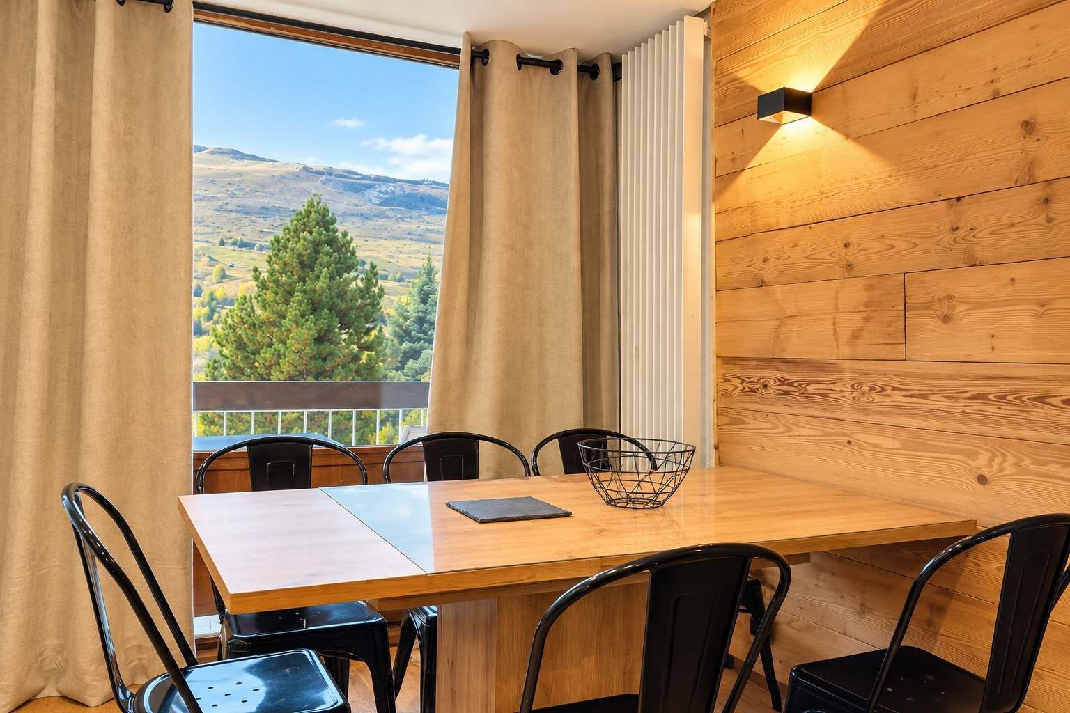 Ein Essbereich mit einem Holztisch, schwarzen Stühlen, einer schwarzen Metallschüssel und Blick auf eine gebirgige Landschaft durch ein Fenster.