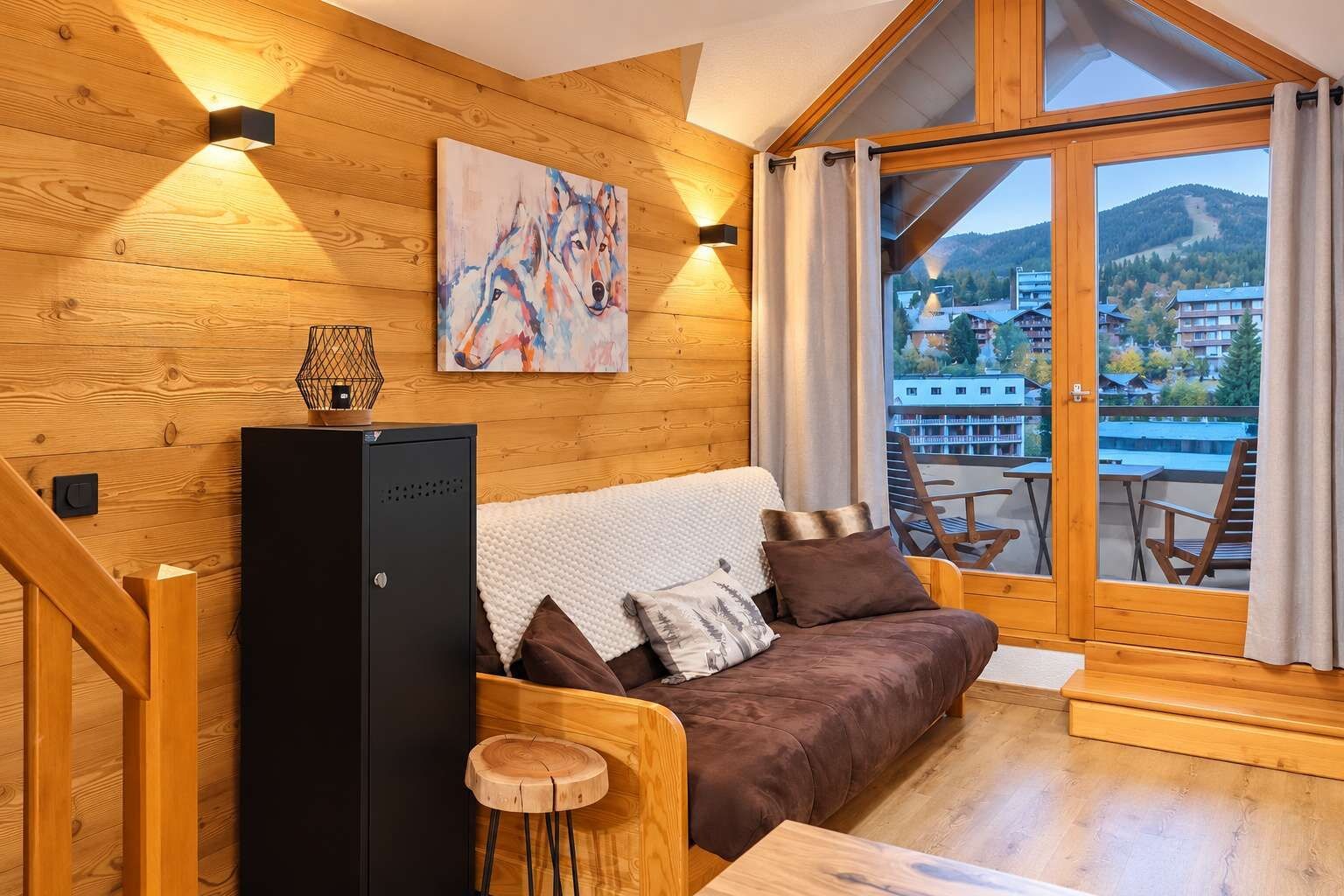 Interior acollidor d'una cabana amb elements de fusta, un sofà amb coixins, una taula petita, un objecte decoratiu a la paret i vista a un paisatge de muntanya a través d'una finestra gran.
