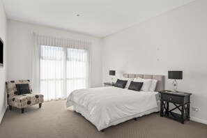 Un dormitorio con una cama, una silla, dos mesitas de noche y un ventanal con cortinas.