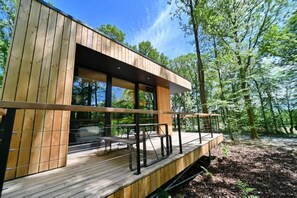 A modern wooden cabin with a deck, large windows, and a view of the forest. - Treehouse Getaway with Sauna (Oignies-en-Thierache)