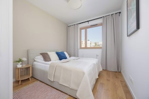 Ein Schlafzimmer mit einem Bett, einem Nachttisch, einem Fenster mit Vorhängen und Blick auf Gebäude.