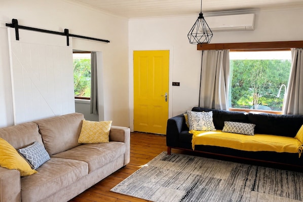 Una sala de estar con un sofá color beige, un sofá negro con cojines amarillos, un tapete estampado, una puerta amarilla y una ventana con cortinas.