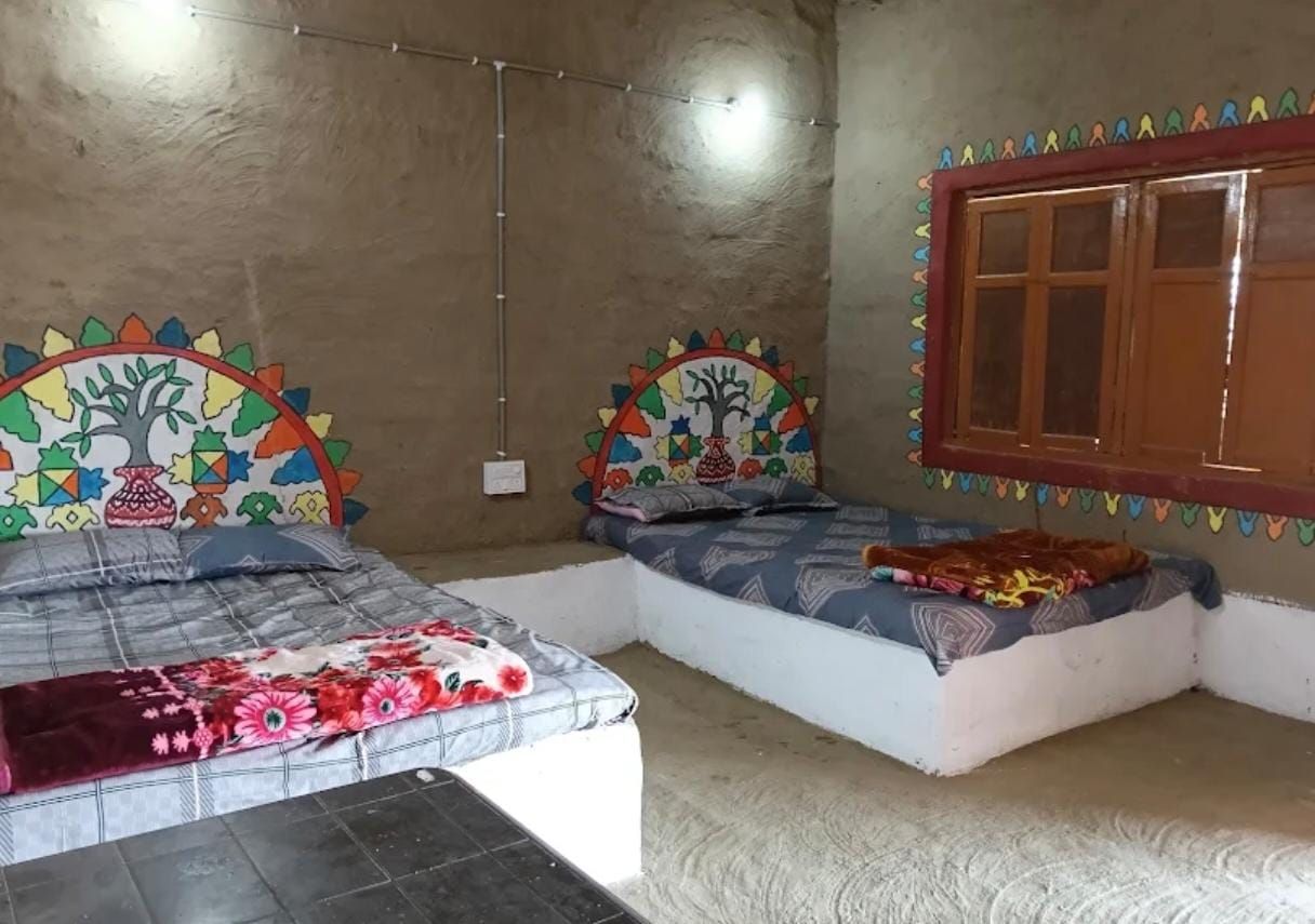 Una habitación con dos camas, una con colcha estampada floral y otra con diseño geométrico. Se ve una ventana con marco de madera y una pared decorada con patrones geométricos coloridos.