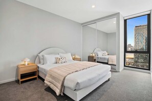 Une chambre moderne avec un grand lit, des tables de chevet et une vue sur le paysage urbain, visible à travers une grande fenêtre.
