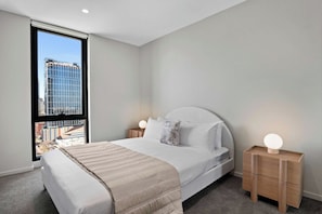 Ein modernes Schlafzimmer mit einem großen Bett, Nachttischen und Blick auf eine Stadtlandschaft durch ein großes Fenster.