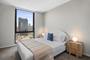 Ein modernes Schlafzimmer mit einem großen Bett, einem Nachttisch mit Lampe und Blick auf eine Stadtlandschaft durch ein Fenster.