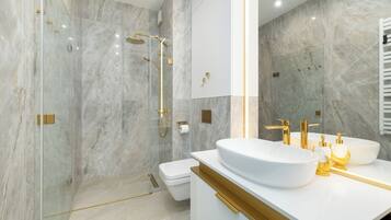 A modern bathroom with a glass-enclosed shower, a white sink with gold fixtures, and a marble wall.