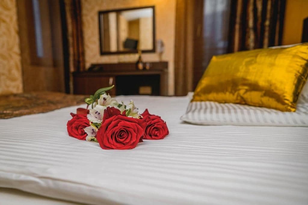 Una cama con sábana blanca, almohada dorada y un ramo de rosas rojas.