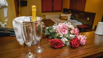 Une chambre d’hôtel avec un lit, une table de chevet, un sèche-cheveux, une bouteille de champagne, deux verres et un bouquet de fleurs.
