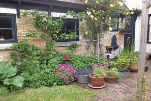 Ein Backsteinhaus mit zahlreichen Fenstern, von denen einige mit außen wachsenden Pflanzen gesäumt sind. Im Vordergrund befinden sich verschiedene Blumentöpfe und Blumen.