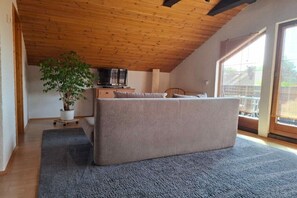 Ein Zimmer mit Holzboden, einem großen Fenster, einer Couch, einem Fernseher und einer Topfpflanze.