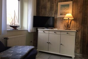 Una habitación con una televisión sobre un mueble, una lámpara, un ventanal con cortinas y un pequeño modelo de velero.