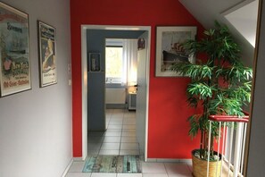 Un couloir avec un mur rouge, une plante en pot et une porte donnant sur une cuisine.