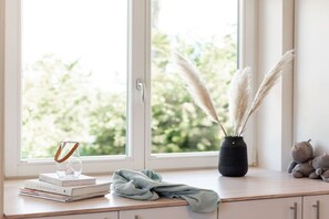 Ein Fenster mit Blick auf Grün, eine Vase mit Pampasgras, ein Stapel Bücher, ein Glas mit Stiel und ein Teddybär.