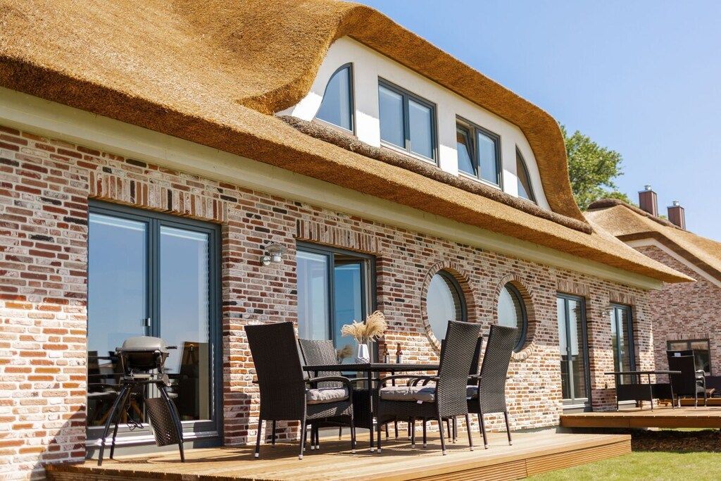 A brick house with a thatched roof, large windows, and an outdoor seating area with chairs and a table.