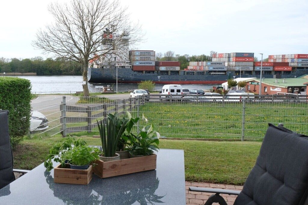 A view of a waterfront area with a large ship docked, a grassy field, and a building with shipping containers.