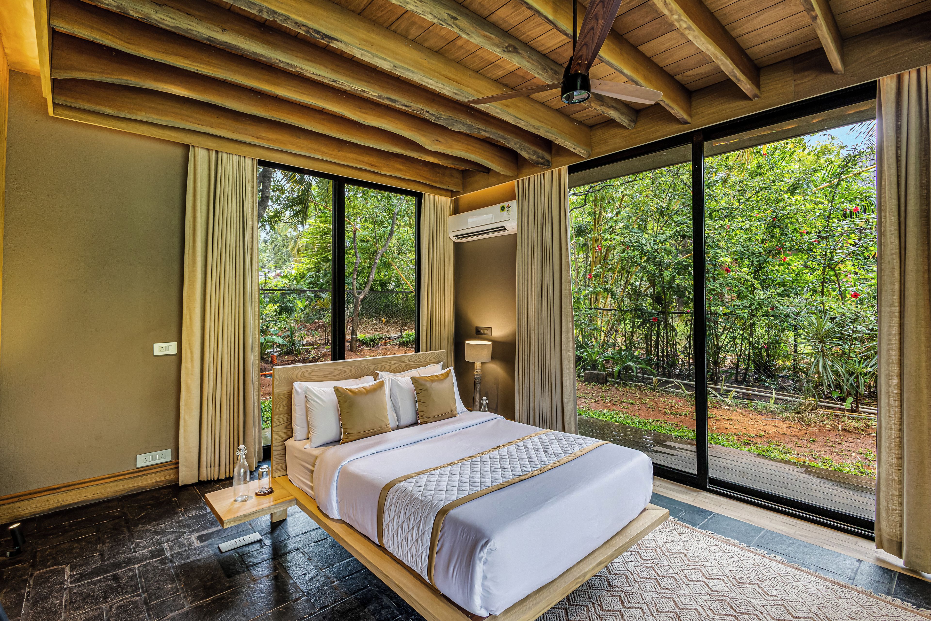 A bedroom with a large bed, a ceiling fan, an air conditioning unit, and a view of a lush garden through large windows.