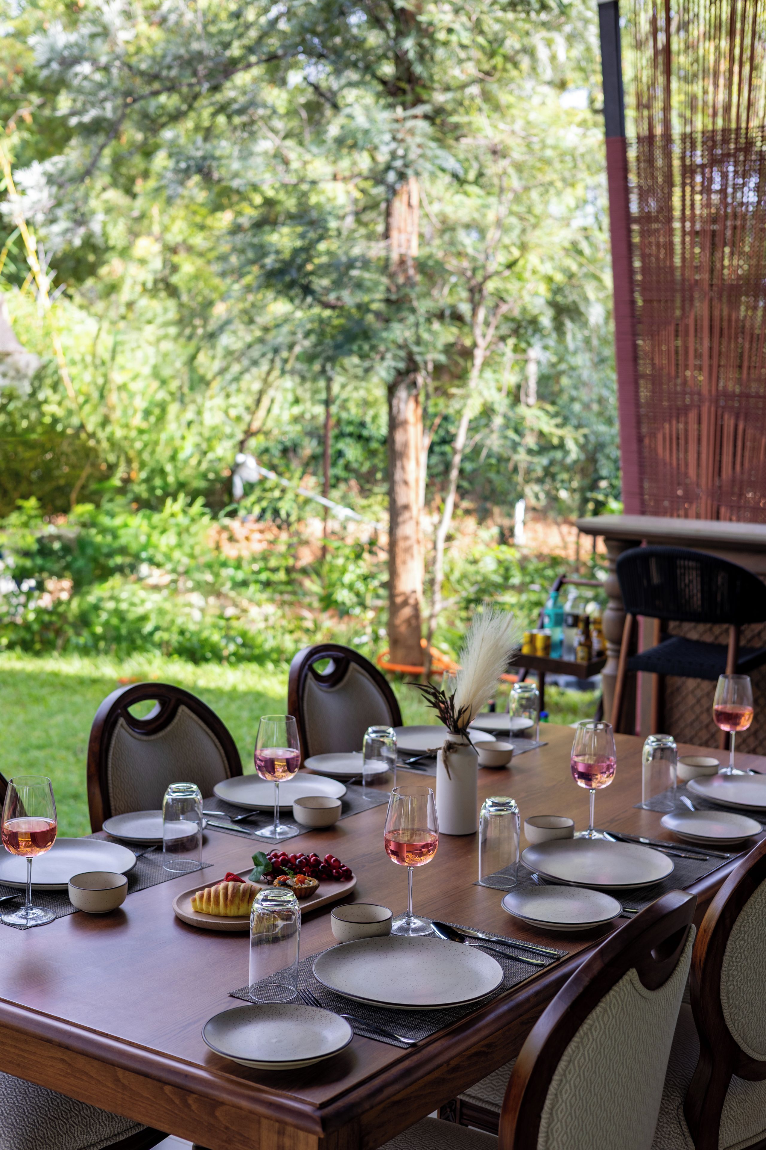 Un área de comedor al aire libre con una mesa dispuesta para una comida, rodeada de sillas y vistas a un jardín exuberante.