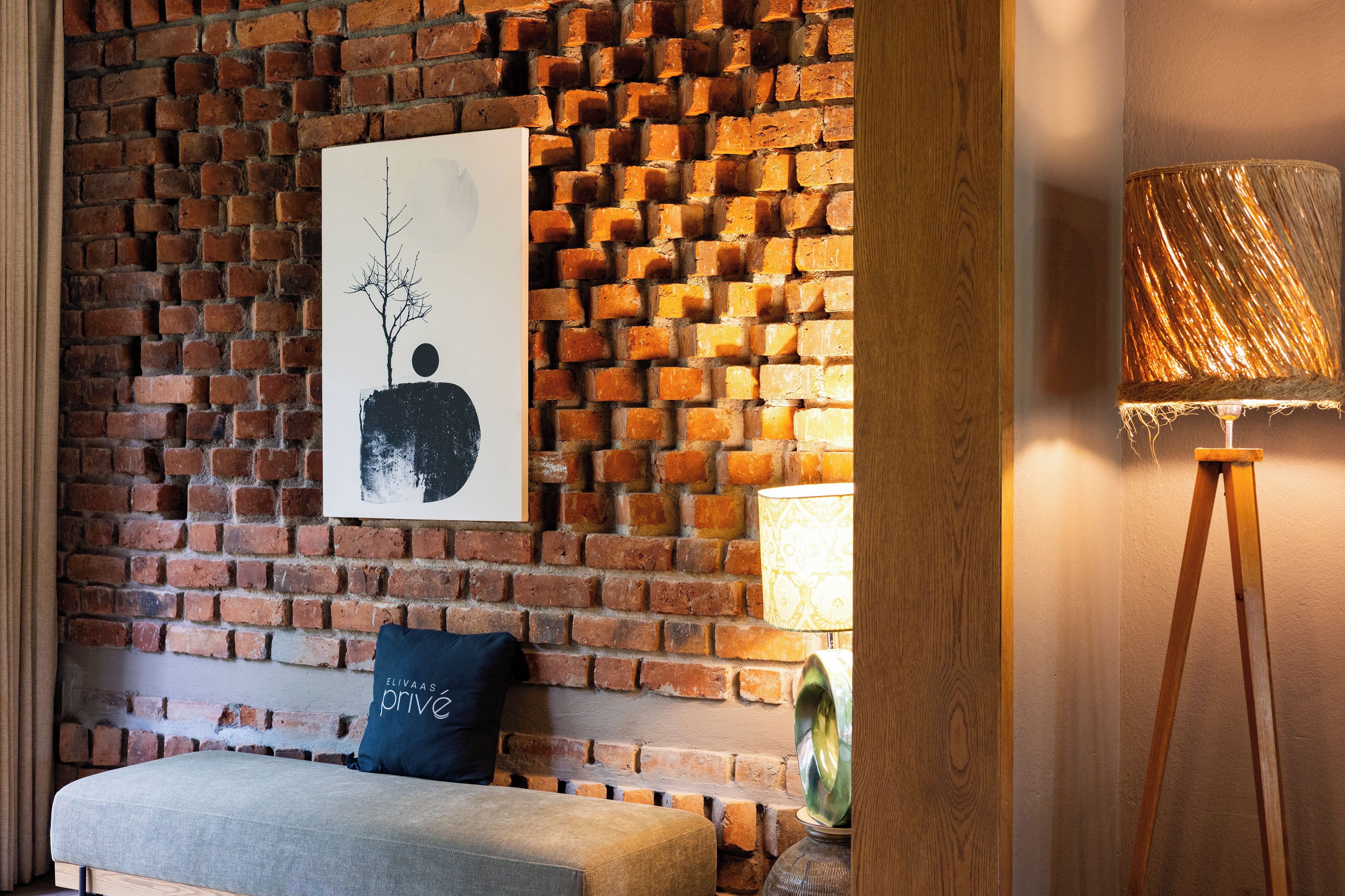 A room with a brick wall, a framed artwork, a lamp, and a bench.