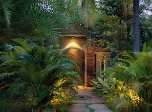 A pathway leading to a brick archway with a light fixture, surrounded by lush greenery.