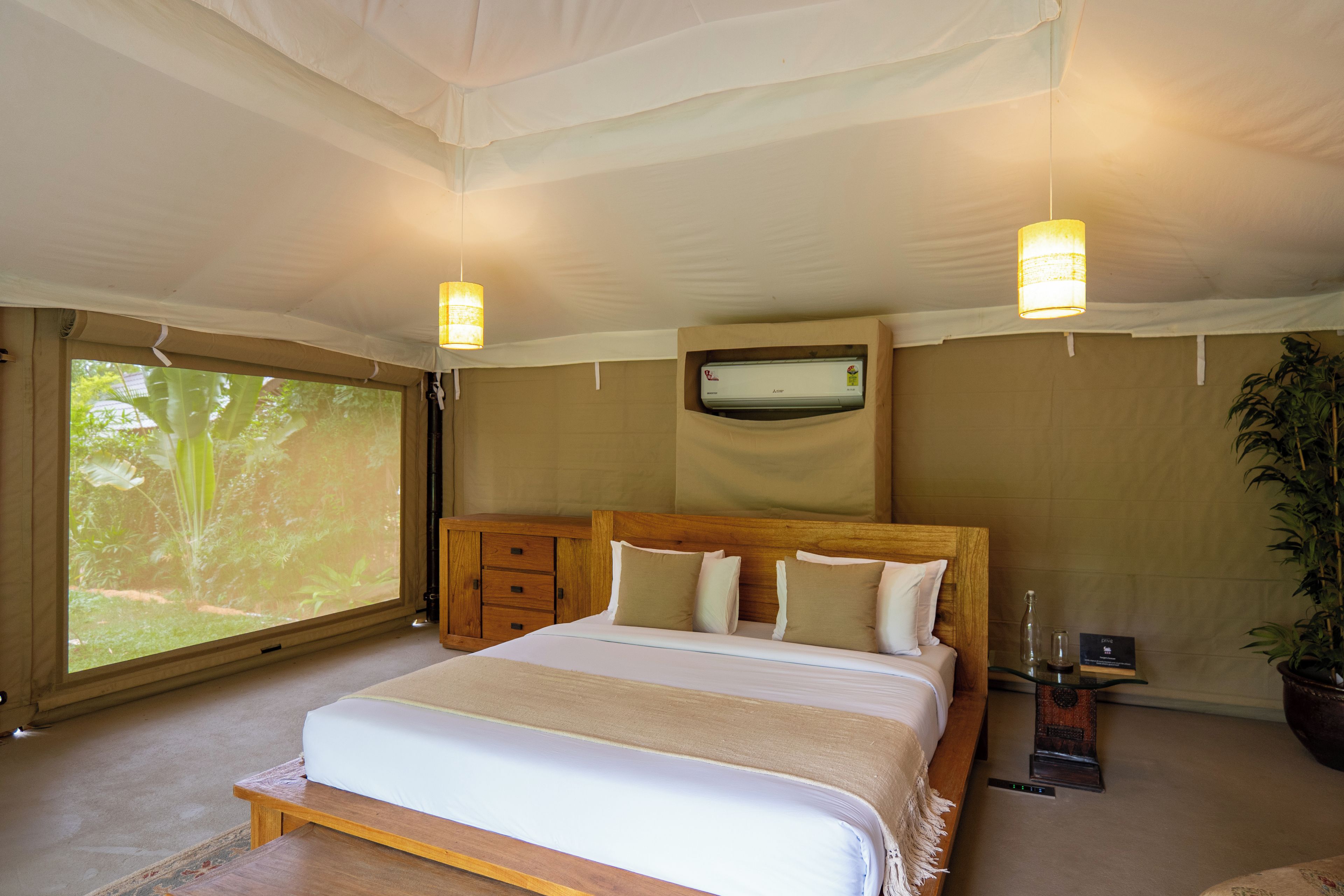 A bedroom with a large bed, a wooden headboard, a flat-screen TV mounted on the wall, a bedside table with a lamp, and a window with a view of greenery.