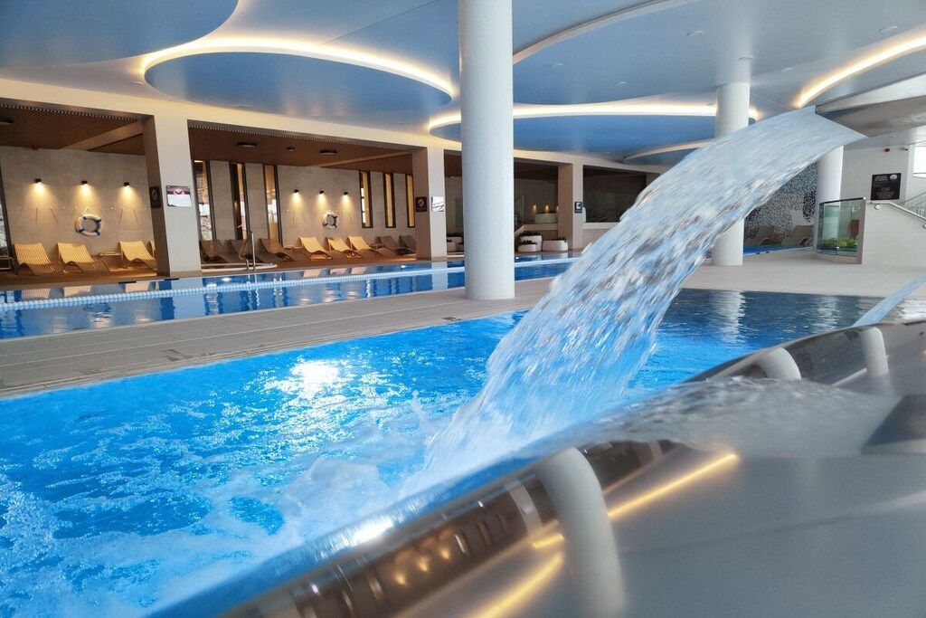 An indoor swimming pool with a water feature, lounge chairs, and a modern architectural design.