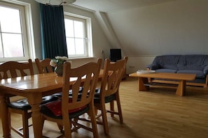 Un área de comedor con mesa y sillas de madera, un sofá y una televisión en una habitación con techos inclinados y ventanas amplias.