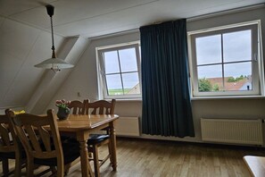 A dining area with a table and chairs, a hanging light fixture, and a view through a window with a curtain. - Holiday apartment with 1 bedroom (Nes Ameland)