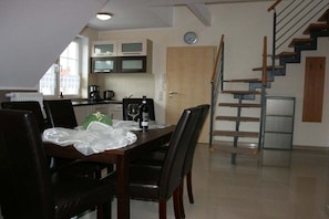 A dining area with a table set for two, a kitchen with cabinets and a microwave, and a staircase with a metal railing. - Villa Guta, Apartment 2 (Rowy  Polen)