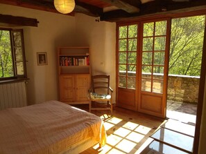 Ein Schlafzimmer mit einem Bett, einem Holzsessel, einem Bücherregal und einem großen Fenster mit Blick auf Bäume.