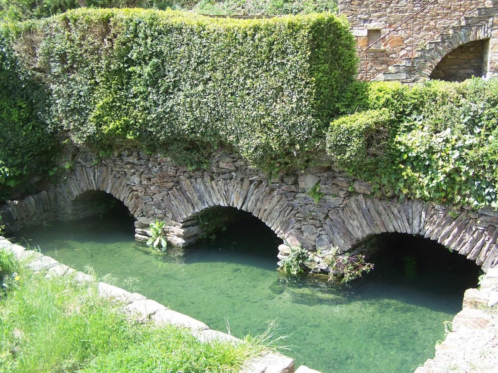 A stone bridge with arches over a clear water channel, surrounded by greenery.