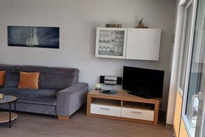 Ett modernt vardagsrum med en grå soffa, en platt-TV på ett träställ, ett glasskåp med dekorativa föremål och en målning av en segelbåt på väggen.