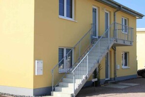 A two-story building with a yellow exterior, a staircase with metal railings, and a small balcony on the upper floor. - Holiday apartment with 1 bedroom (Gelbensande)