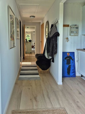 Un couloir avec un plancher en bois, un fauteuil noir à la texture duveteuse, un porte-manteau et un tableau accroché au mur.